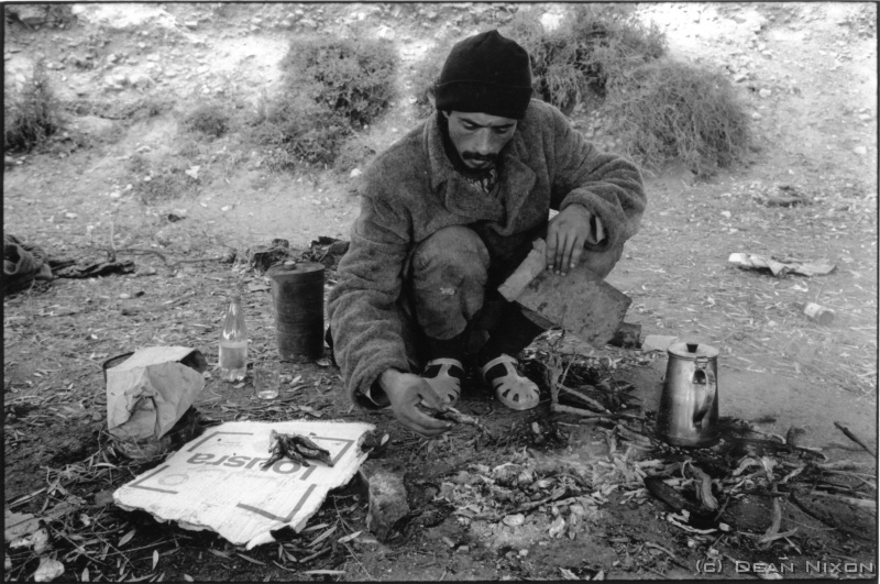 2001.02 Morocco. Homeless Man_hf <div align=left>Feb 2001<br>"Morocco was a blast" A short-list of impressions: A Tuareg (tough guys of the Sahara) orders a schoolboy off his bike and rides away. A German woman saunters topless onto a public beach only to be roughly and rapidly hauled away in tears by two burly policemen. Street peddlers shout "Chocolate? Hashish?..." while a shopkeeper doesn t even look up from his Koran. A homeless man invites me for sweet tea and BBQd sardines, and washes my hands with bottled water. A hand-drawn cart is passed in a cloud of dust by a new Mercedes. The Levis and Calvin Kleins have no button holes. I play football in an alley with laughing children in rags. Tourists clutch their bags nervously and scurry past. Traditionally dressed women avoid pointed cameras instantaneously while young girls make a point of flashing their eyes at foreigners. A row of shoe-shine boys with boxes of Kiwi brand polish while a man sells guns outside a bank. <br>Old cultures meet new with an odd harmony here...mostly. A deformed old man is quietly begging opposite a mosque...passing locals give him a few coins. A group of overweight Europeans pause beside him, one standing on his bench to get a better camera angle. He stretches his hand towards them, but without a glance they amble away. "Wirklich Scheie" I thought. As I left he said something softly about Allah.<br>